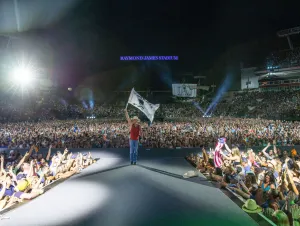 Kenny Chesney Found More Tickets - Kenny is wearing a red shirt and cowboy hat, waving a flag while playing Tampa's Raymond James Stadium in 2022.