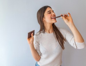 Young woman with natural make up having fun and eating chocolate isolated on gray background. Woman eating chocolate. Happy teenage girl eating chocolate bar