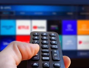 A man is holding a remote control of a smart TV in his hand. In the background you can see the television screen with streaming entertainment apps for streaming Hulu, Max, Disney+