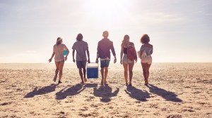 Florida destinations for Memorial Day. Rear view portrait of group of friends walking on the beach and helping each other while carrying a cooler box.