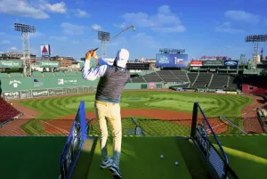 Upper Deck Golf at Fenway Park. A golfer takes a swing inside the baseball park on a sunny day.