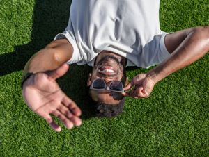 An African-American man in his 30s wearing sunglasses lies sprawled on the grass of a well-maintained public park.