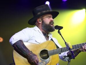 Zac Brown is wearing a white shirt and hat on stage singing.