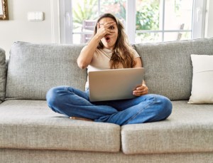 oung brunette woman sitting on the sofa using computer laptop at home peeking in shock covering face and eyes with hand, looking through fingers with embarrassed expression