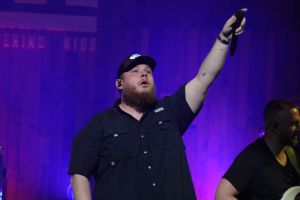 Luke Combs wearing a black button down shirt and a black cap on stage holding a mic