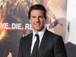 Tom Cruise wearing a suit in front of an "Edge Of Tomorrow" movie poster.