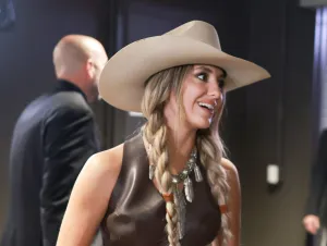 Lainey Wilson is smiling in a brown suit, ponytails, and cowboy hat.