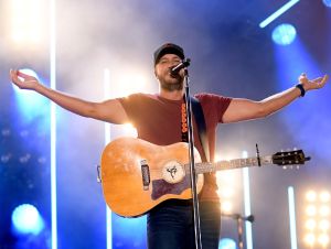 Luke Bryan is on stage in a bronze t-shirt and a guitar.